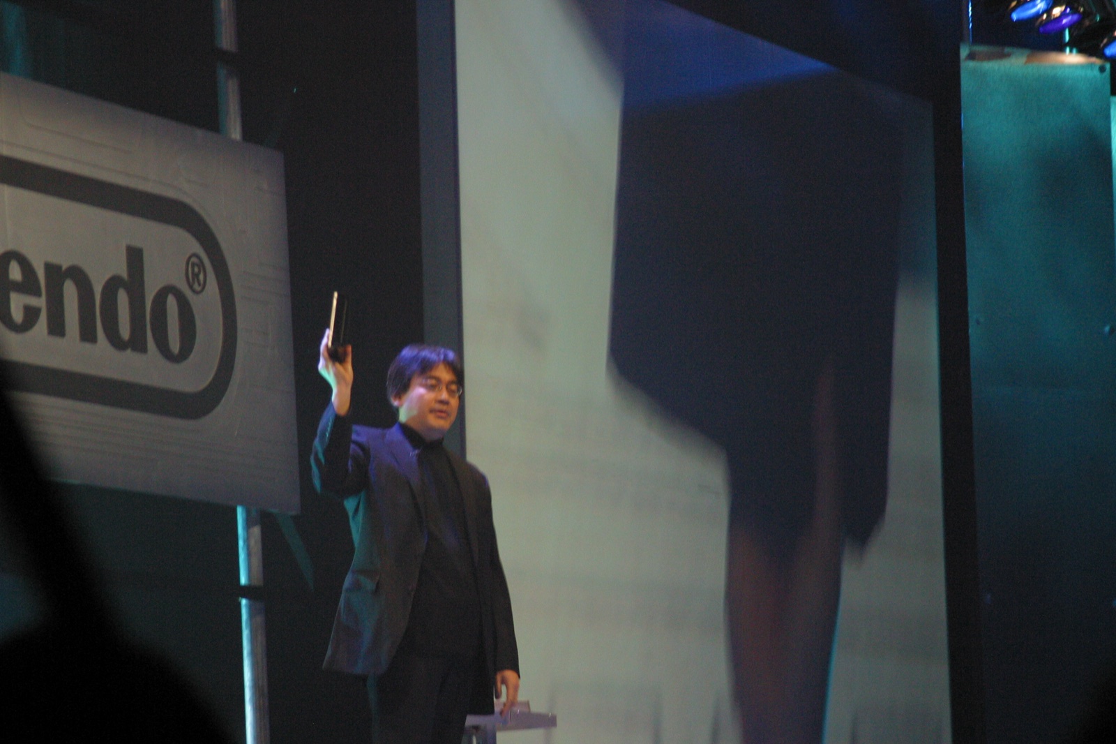 Satoru Iwata on stage at the Nintendo press conference, holding up a Revolution (soon to be called Wii) for the audience.