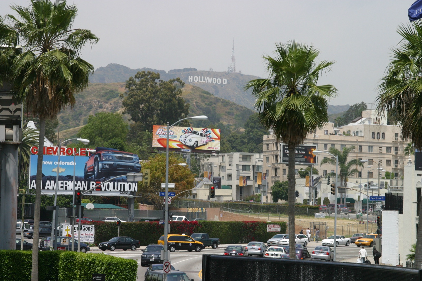 The Hollywood sign in the haze, framed by palm trees and billboards — the view from LA.